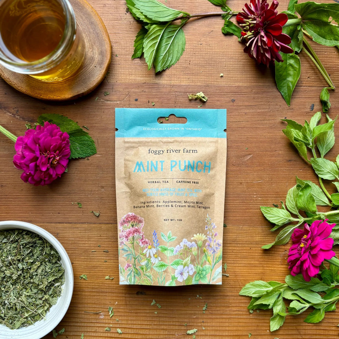 Tea package labeled 'Mint Punch' from Foggy River Farm on a wooden surface with tea leaves and flowers.