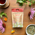 Tea package labeled 'Boost' surrounded by flowers and tea leaves on a wooden surface