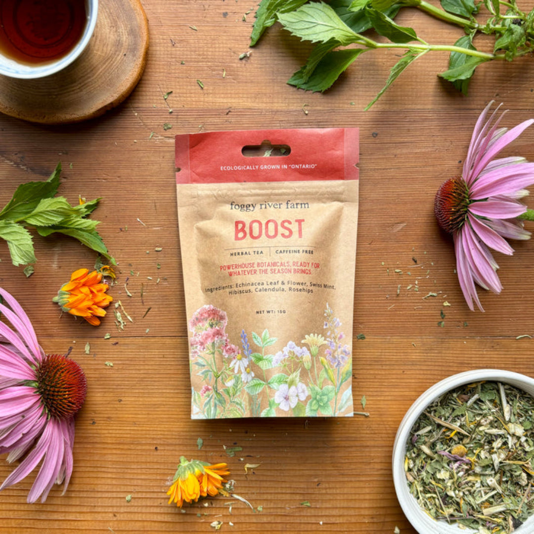 Tea package labeled 'Boost' surrounded by flowers and tea leaves on a wooden surface