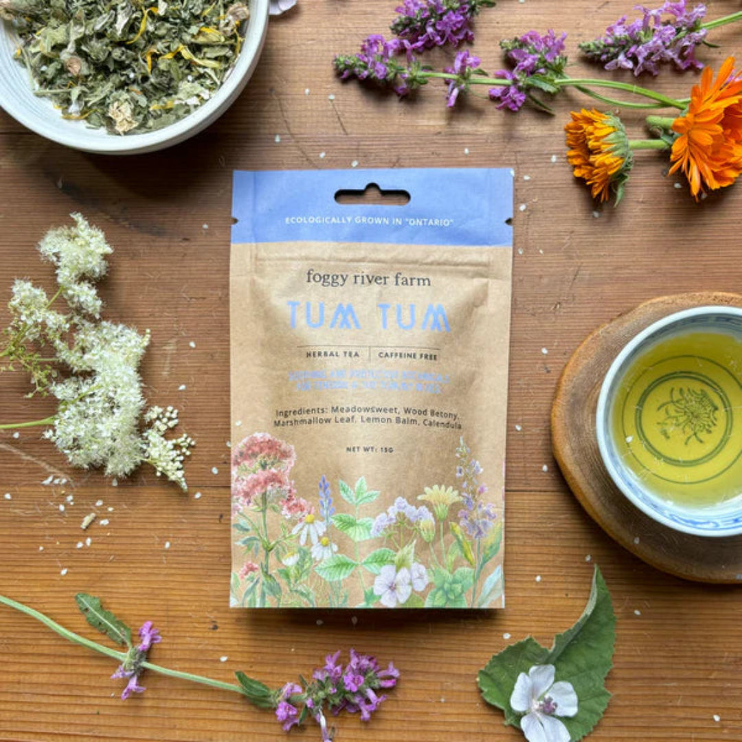 Foggy River Farm Tum Tum herbal tea package on a wooden surface with flowers and tea.
